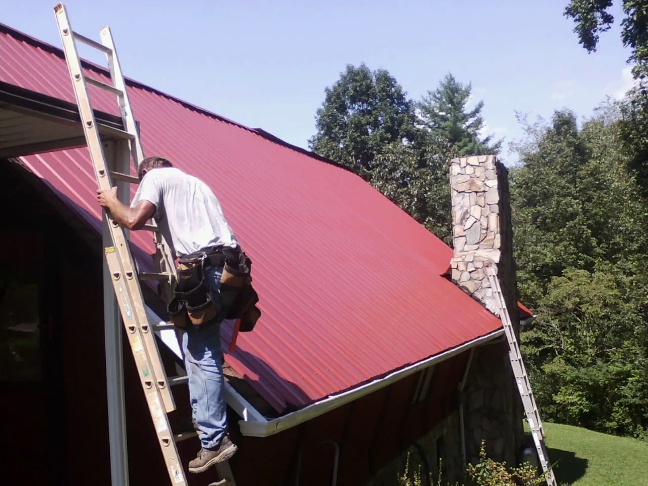 Roofer on a ladder beside a red metal roof and stone chimney — Soffit & Fascia Repair by Roofing Brothers in East Wakefield